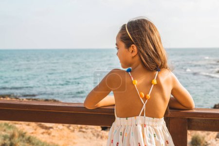 Photo for Back view of a beautiful little girl in summer dress looks at the sea. - Royalty Free Image