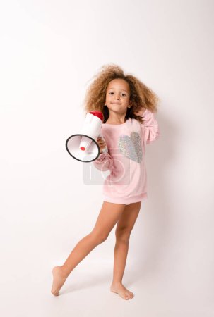 Téléchargez les photos : Petite fille afro-américaine de 4 ans en robe décontractée tenant un mégaphone isolé sur fond blanc portrait d'enfant en studio. Concept de style de vie des enfants - en image libre de droit