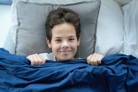Photo for Smiling preteen boy child lying in bed and covering his tricky playful face with a blanket - Royalty Free Image