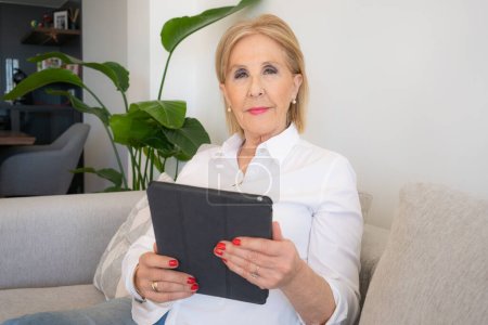 lächelnde ältere Frau mit digitalem Tablet entspannt auf Sofa sitzen und E-Book in Leser-App lesen, glückliche Senioren-Oma mittleren Alters beim Online-Shopping auf dem PC zu Hause