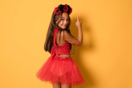 Cute smiling little kid girl in red halloween costume pointing finger up over orange background. Halloween concept.