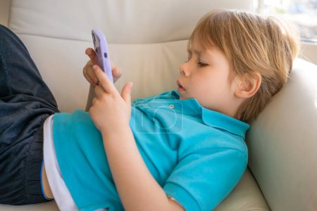 Nahaufnahme niedlicher kleiner Junge mit Smartphone, Blick auf Bildschirm, neugieriges Kind, das Handy in der Hand hält, allein zu Hause auf der Couch sitzt, Handyspiele spielt, Cartoons online anschaut.