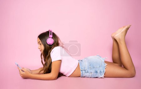 Foto de Niña sentada en el suelo, usando un smartphone y escuchando la música. Vista lateral. Estudio de larga duración tiro aislado en rosa, - Imagen libre de derechos