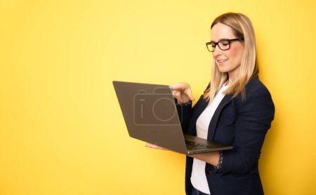 Téléchargez les photos : Souriant joyeux drôle belle jolie jeune femme 40s portant veste décontractée de base debout travaillant sur ordinateur portable PC isolé sur fond de couleur jaune portrait studio - en image libre de droit