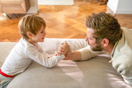 Photo for Raising a real man. Concentrated father and son arm wrestling and competing while lying on floor, having fun at home, side view. Family bond, leisure time and fatherhood concept - Royalty Free Image