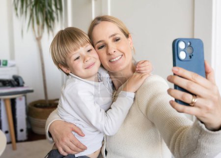 Lächelnde junge Mutter und kleiner Sohn, die zusammen telefonieren, sich umarmen, zu Hause, glückliche Mutter mit entzückendem Jungen, der ein Selfie macht, Freizeit mit Gadget genießt