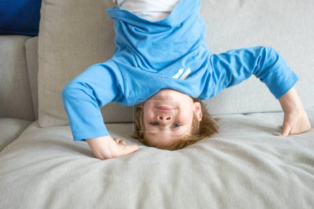 Cute funny boy lying upside down on sofa looking at camera, smiling playful preschool child having fun at home on couch