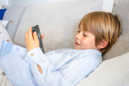 Niño lindo sonriendo usando teléfono inteligente tumbado en el sofá en casa