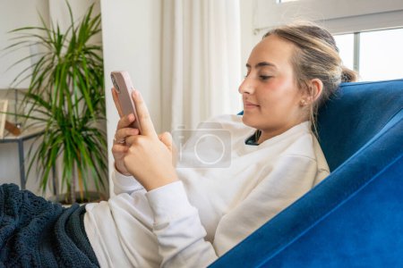 lächelnde Teenagermädchen entspannen auf dem Sofa zu Hause sitzen und mit Freunden per Handy-SMS chatten, glückliche junge Frau auf der Couch Smartphone-Shop online halten oder mobile Anwendung oder soziale Medien checken