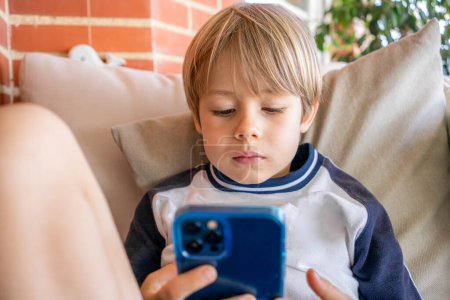 Kleiner lustiger Junge spielt Smartphone-Spiel während er zu Hause auf dem Sofa sitzt