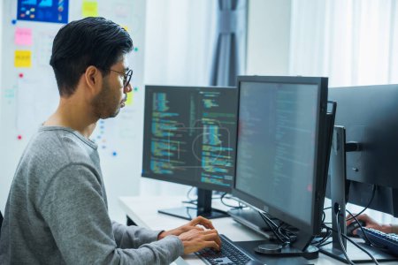 Téléchargez les photos : Asiatique homme invite ingénieur développer application de codage avec des données logicielles assis devant moniteur d'ordinateur au bureau - en image libre de droit
