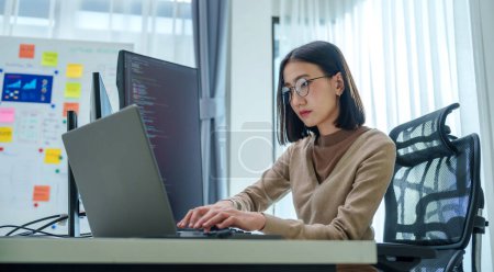 Téléchargez les photos : Ingénieur invite asiatique développer application de codage avec des données logicielles assis devant le moniteur d'ordinateur au bureau - en image libre de droit