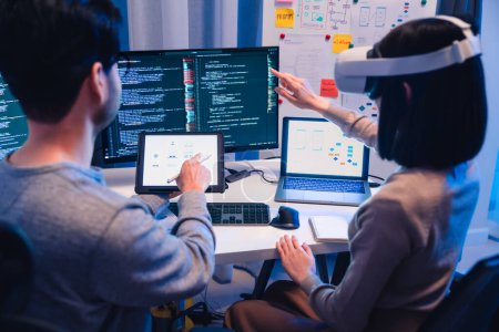 Téléchargez les photos : Développeur de logiciels asiatiques portant un casque de réalité virtuelle travaille sur un projet VR, avec une collègue axée sur son ordinateur - en image libre de droit