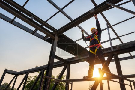 Téléchargez les photos : Un travailleur de la construction travaillant en hauteur à l'aide du crochet pour harnais de sécurité sur la structure du toit sur le chantier de construction. - en image libre de droit