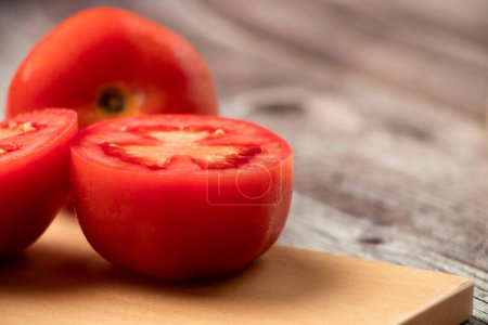 Foto de Un tomate a la mitad en una tabla de cortar. Todo se encuentra sobre una mesa gris rstica. Cerca de la mesa hay otros tomates. Fotografa de primer plano. - Imagen libre de derechos