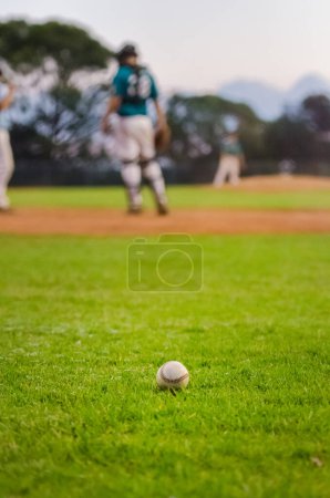 Foto de Pelota de béisbol sentada en el campo de diamantes de hierba. Catcher y lanzador en el fondo borroso. - Imagen libre de derechos
