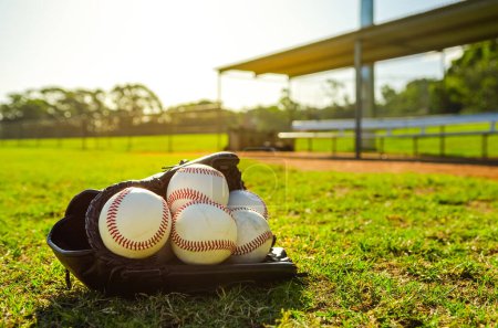 Foto de Guante de béisbol lleno de bolas de béisbol sentado en el campo de pelota en frente de dugout vacío durante una puesta de sol - Imagen libre de derechos
