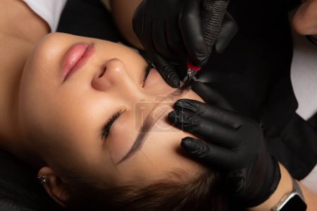 Cosmetician in black gloves making permanent makeup to a woman with a markup on a brows