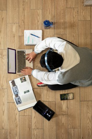 Photo for Young man with headphones sitting on the floor of his house studying and working at home with a computer, tablet, book, cell phone and glass of water. - Royalty Free Image