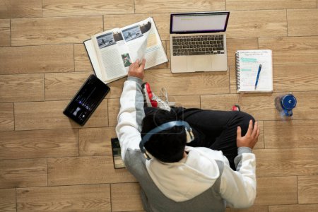 Young man with headphones sitting on the floor of his house studying and working at home with a computer, tablet, book, cell phone and glass of water.