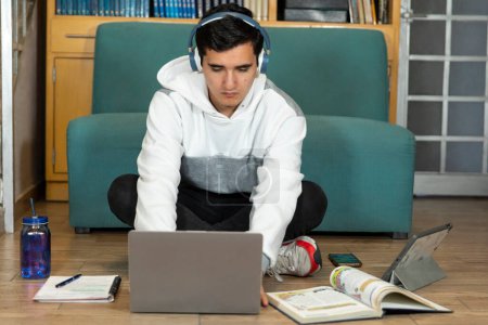 Photo for Young man with headphones sitting on the floor of his house studying and working at home with a computer, tablet, book, cell phone and glass of water. - Royalty Free Image