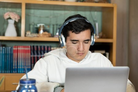 Photo for Young man with headphones sitting on the chair of his house studying and working at home with a computer, tablet, book, cell phone and glass of water. Home work. - Royalty Free Image