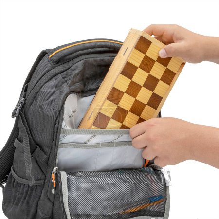 Foto de Man's hand putting into backpack. Traditional handmade wooden chess board with black and natural colored chips on a white background. - Imagen libre de derechos