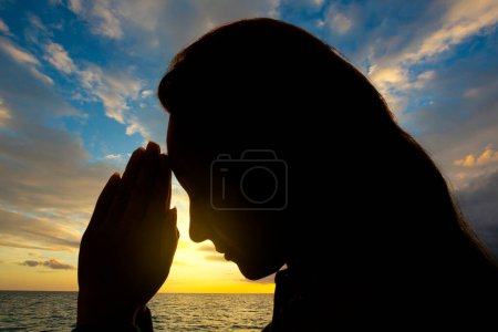 Woman's face backlit with hands in prayer on a sunset background.