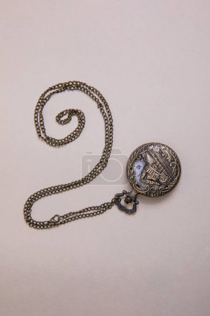 antique pocket watch with train engraving on the front cover, patinated brobce color, chain arranged in a spiral on a beige background overhead view