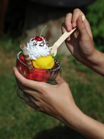 Tasting ice cream sundae with three kinds of ice cream, whipped cream, cherry with a wooden spoon in nature, hot summer day, concept without plastic, young women hands
