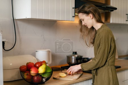 Téléchargez les photos : Une jeune femme caucasienne en robe kaki coupe une pomme dans une cuisine moderne. Concept de saine alimentation - en image libre de droit