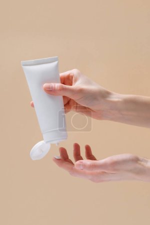 Vertikales Bild weiblicher Hände, die Creme aus einem leeren Rohr auf einem Finger auf einem beigen isolierten Hintergrund auspressen. Das Konzept der Schönheit und Ästhetik. Image für Ihr Design