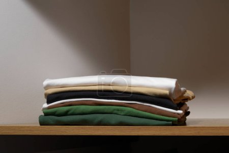 Photo for Folded t-shirts of white, beige and green colors on a wooden shelf of a closet. The concept of compact storage of clothes, changing or updating the wardrobe - Royalty Free Image