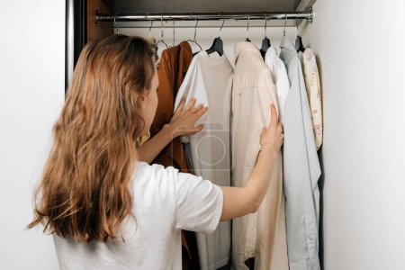 Eine junge kaukasische Frau wählt Hemden und T-Shirts aus einem Kleiderschrank, der an Kleiderbügeln in einem Kleiderschrank hängt. Das Konzept, die Garderobe vom Sommer auf den Winter zu wechseln, neue Kleidung zu kaufen
