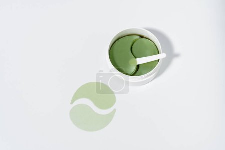 Téléchargez les photos : Pot avec taches d'hydrogel vert et spatule sur fond blanc isolé. Le concept de produits pour la peau sous les yeux, soin du visage - en image libre de droit