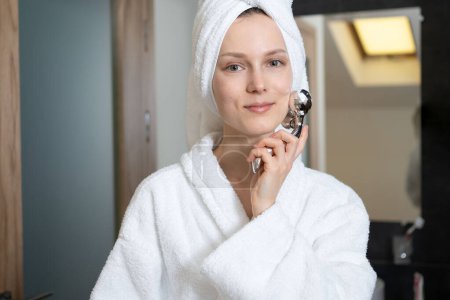 Téléchargez les photos : Une jeune femme avec une serviette sur les cheveux et un peignoir se masse le visage avec un masseur miroir. Soins du visage, produits de beauté et concept de spa - en image libre de droit