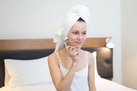 Téléchargez les photos : Une jeune femme avec une serviette sur les cheveux se masse le visage avec un masseur miroir 3D. Le concept de lifting et soin du visage, produits de beauté - en image libre de droit