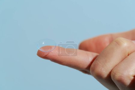 Finger der weiblichen Hand hält eine blaue transparente Kontaktlinse, während sie auf blauem, isoliertem Hintergrund getragen wird. Sehverbesserungskonzept, Weit- und Kurzsichtigkeit, Augenarzt.