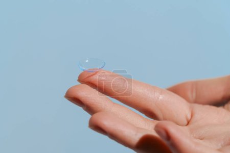 Finger of female hand holding transparent contact lens on blue isolated background. Concept of ophthalmology, improvement of vision