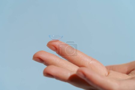 mano femenina sosteniendo lente de contacto azul claro antes de poner en el ojo sobre fondo aislado azul. Concepto de mejora de la visión, miopía.
