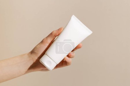 Mano femenina sosteniendo una maqueta de tubo blanco sobre fondo aislado beige. Concepto de belleza, estética, productos de cuidado de la piel.