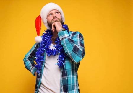 Téléchargez les photos : Attrayant jeune homme barbu dans une chemise à carreaux portant un chapeau de Père Noël et avec un boa bleu avec un visage réfléchi sur un fond jaune - en image libre de droit