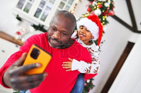 Foto de Padre en un suéter rojo con un hijo pequeño en sus hombros en un sombrero de santa sonriendo mientras se filmaba en una cámara de teléfono inteligente sobre el fondo de un árbol de Navidad. - Imagen libre de derechos