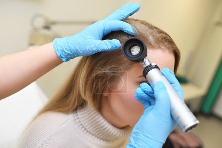 A dermatologist trichologist examines the hair structure of a young womans patient using an optical dermatoscope device.