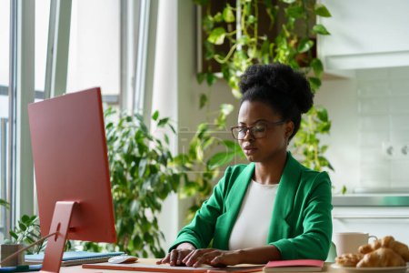 Erfolgreiche Afroamerikanerin, die als Redakteurin eines Wirtschaftsmagazins arbeitet und Artikel tippt, sitzt am Computertisch. Mädchen mit Brille verrichtet Fernarbeit für Internetpresse im Homeoffice mit Pflanzen