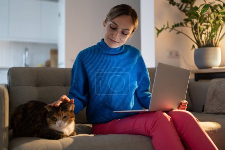 Female freelancer wearing blue sweater sits on sofa stroking domestic cat at sunset. Lonely woman holding notebook on laps admires cat and enjoys having pet friend in cozy apartment with potplants