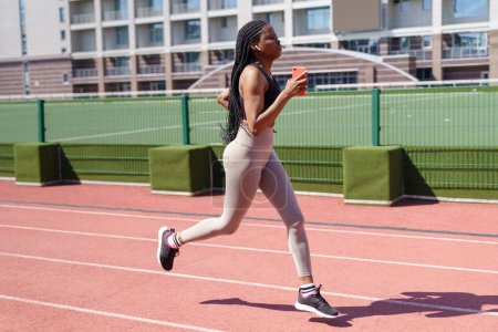 Photo for African American female enjoys running every day on red track listening to music in headphones on hot summer day. Young woman tries to keep healthy lifestyle at sports ground of residential complex - Royalty Free Image