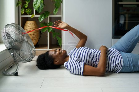 Happy young African woman lying on floor enjoying air flow from electric portable fan, cooling off house without air conditioning in heat. Satisfied black female resting at home during summer day