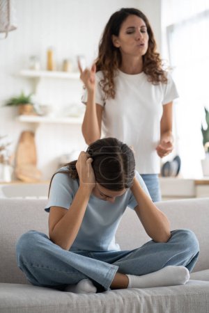 Photo for Sad teenage girl sits cross-legged on couch and covers ears with hands not wanting to listen to mother reproaches. Nervous woman looking at daughter scolding after teacher call from school. Parenting. - Royalty Free Image