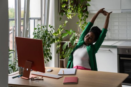 Femme africaine fatiguée freelance faire des exercices d'étirement, travaillant à distance sur ordinateur dans la cuisine à la maison. Fille noire prenant une pause de travail indépendant ou d'étude en ligne, se détendre au travail à distance
